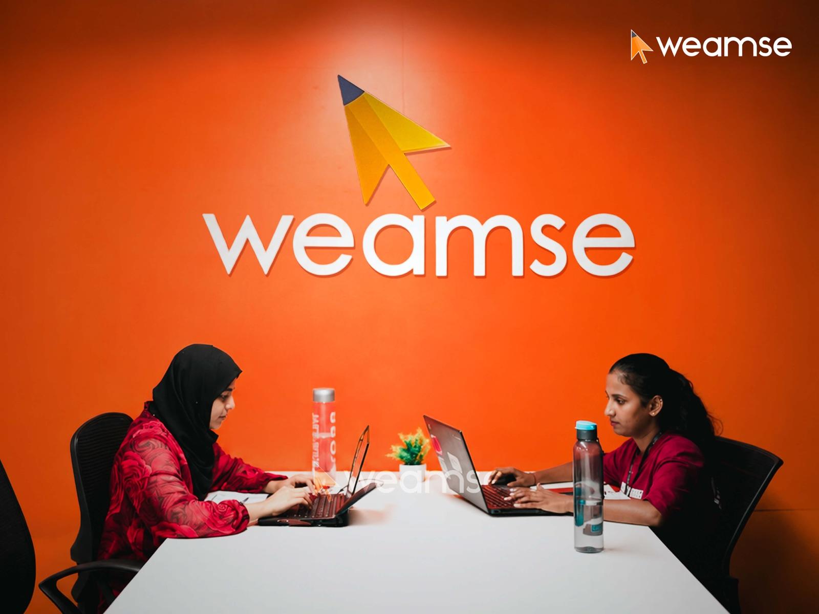 Two team members at work beneath the WEAMSE logo wall